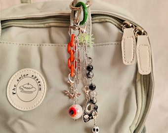 May include: A collection of spooky-themed charms and chains, including a bat, eyeball, and skull, hang from a green carabiner clip. The charms are attached to a light green backpack. The charms are orange, silver, black, and white.