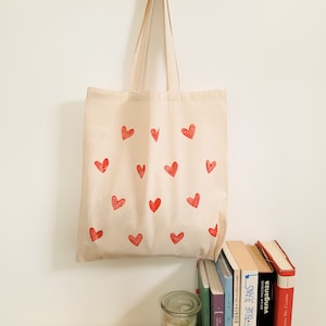 May include: A natural canvas tote bag with red heart designs printed on the front. The bag has a natural canvas strap.
