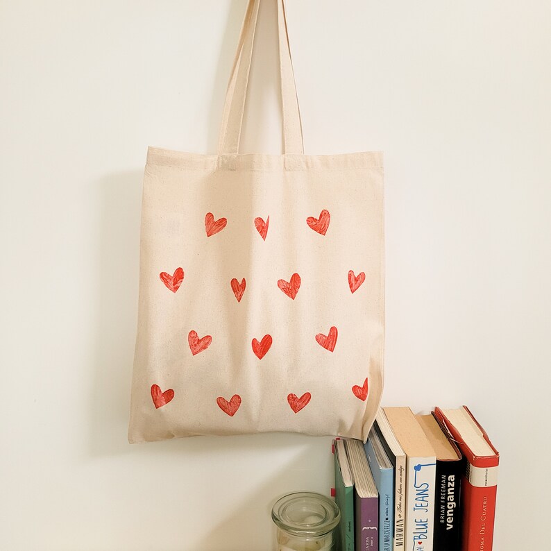 May include: A natural canvas tote bag with red heart designs printed on the front. The bag has a natural canvas strap.