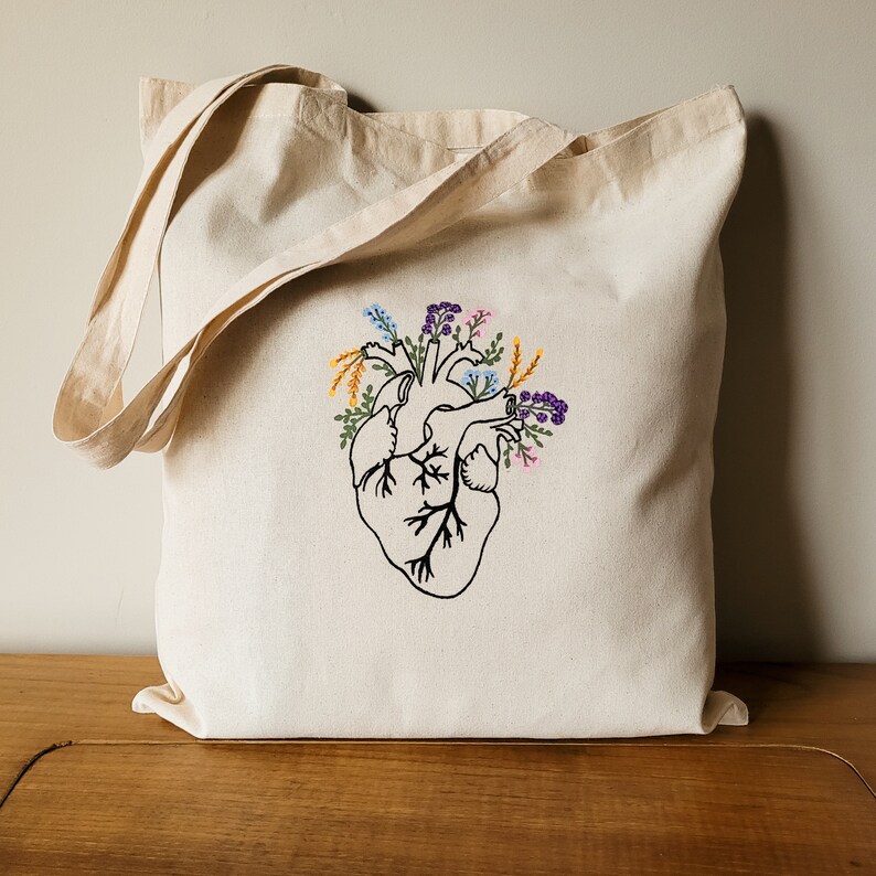 K&ouml;nnte beinhalten: Eine wei&szlig;e Canvas-Tragetasche mit einem schwarzen gestickten anatomischen Herzdesign. Das Herz ist von bunten Blumen in Blaut&ouml;nen, Lila, Gelb und Orange umgeben.