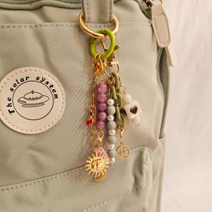 May include: A collection of decorative charms and beads attached to a gold-colored keyring. The charms include a sun and moon pendant, a heart-shaped charm, and various beaded strands in shades of pink, white, and green. The keyring is attached to a light green backpack.