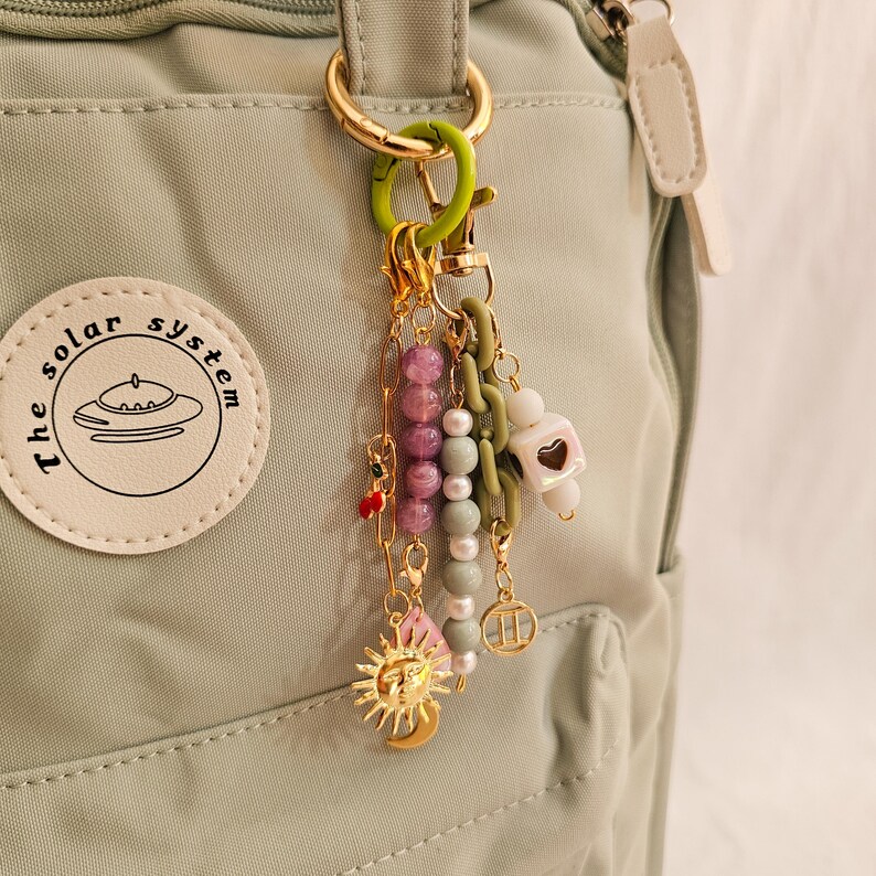 May include: A collection of decorative charms and beads attached to a gold-colored keyring. The charms include a sun and moon pendant, a heart-shaped charm, and various beaded strands in shades of pink, white, and green. The keyring is attached to a light green backpack.