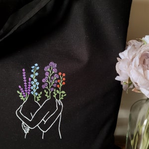 May include: A black tote bag with a white outline of two figures embracing. The figures have flowers growing from their heads, including purple, blue, and orange flowers.