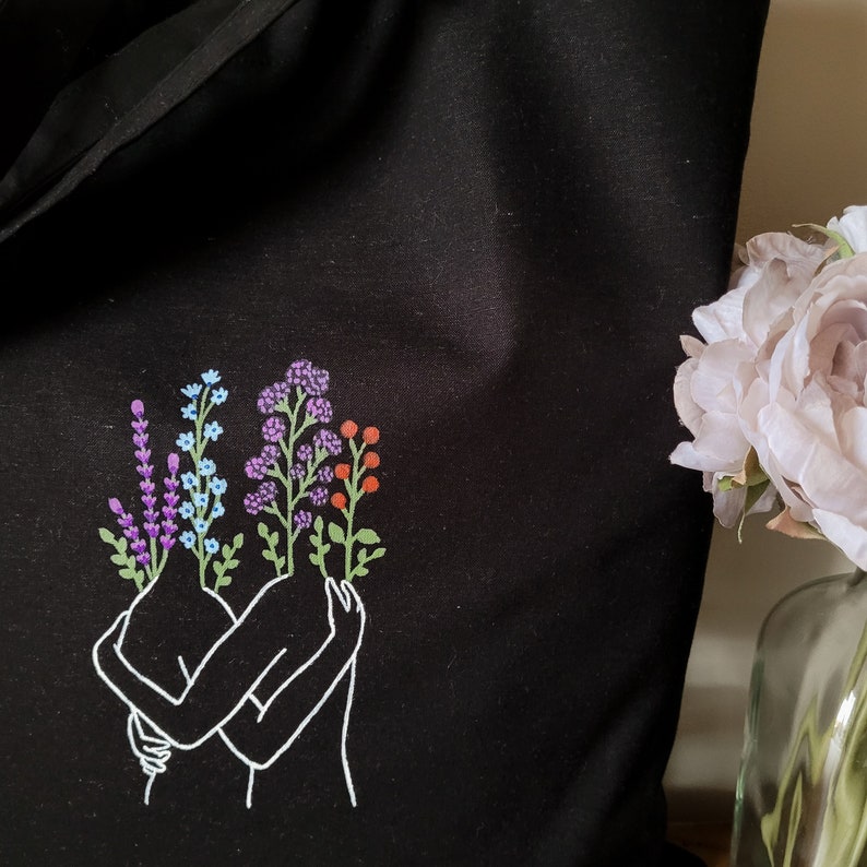 May include: A black tote bag with a white outline of two figures embracing. The figures have flowers growing from their heads, including purple, blue, and orange flowers.
