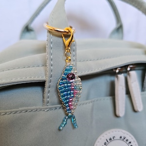 May include: A beaded fish charm with blue, pink, and white beads, a purple eye, and gold-colored hardware. The charm is attached to a gold-colored ring and clasp, hanging from a light green backpack.