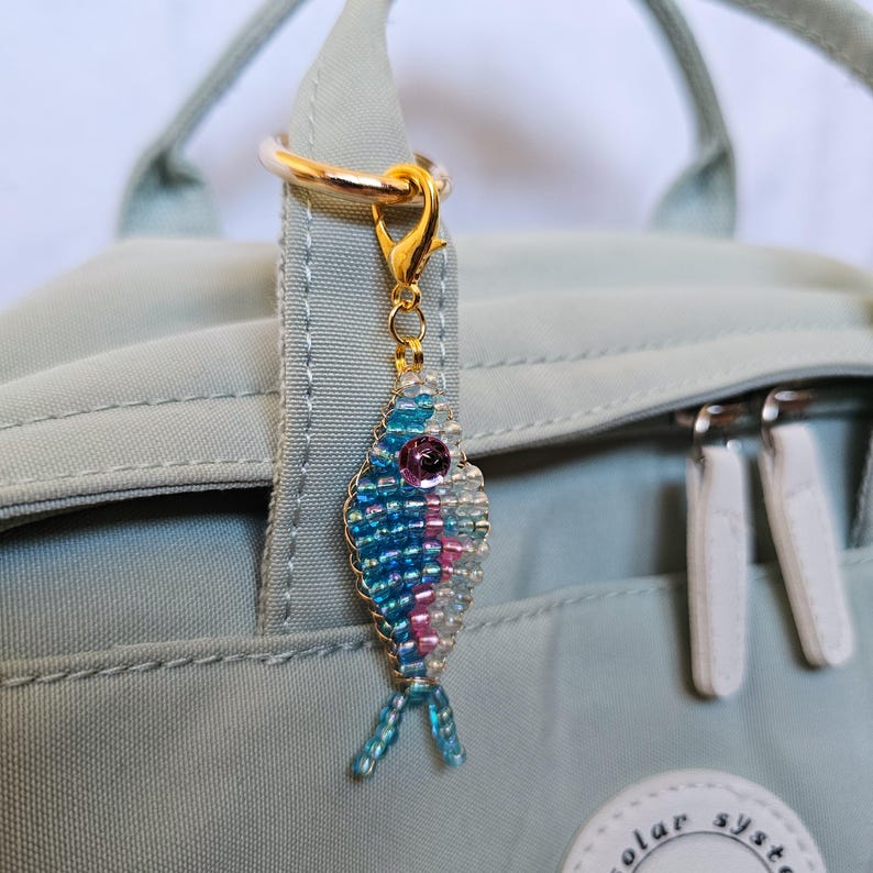 May include: A beaded fish charm with blue, pink, and white beads, a purple eye, and gold-colored hardware. The charm is attached to a gold-colored ring and clasp, hanging from a light green backpack.