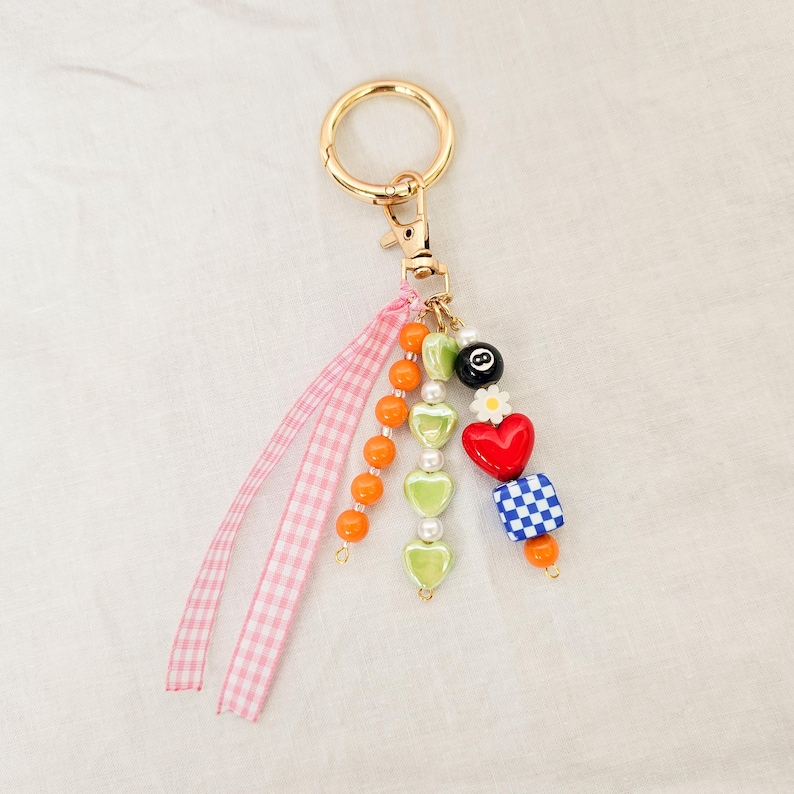 May include: A gold-colored keychain with a variety of colorful beads and charms. The charms include orange and green beads, a red heart, a blue and white checkered square, and a black 8-ball. A pink gingham ribbon adds a decorative touch.