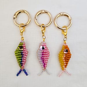 May include: Three beaded fish keychains with gold-colored clasps. The fish are in shades of pink, green, yellow, and orange, with small bead eyes and tail fins. The keychains are on a white background.