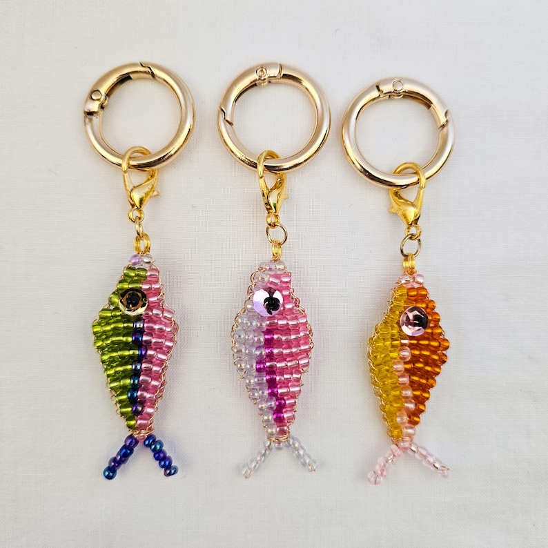 May include: Three beaded fish keychains with gold-colored clasps. The fish are in shades of pink, green, yellow, and orange, with small bead eyes and tail fins. The keychains are on a white background.