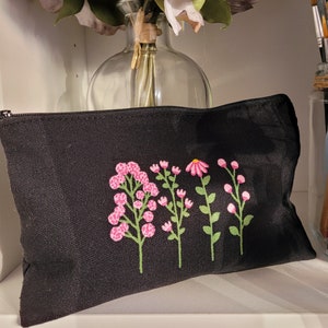 May include: Black canvas zippered pouch with a floral design embroidered in pink. The design features five flowers with pink petals and green stems.
