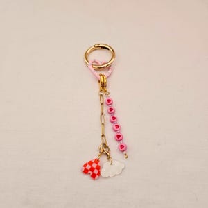 May include: A gold-colored keychain with a pink heart-shaped clasp and a chain. The chain holds a strand of pink heart-shaped beads and two charms: a red and white checkered heart and a white cloud.