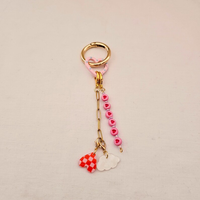 May include: A gold-colored keychain with a pink heart-shaped clasp and a chain. The chain holds a strand of pink heart-shaped beads and two charms: a red and white checkered heart and a white cloud.