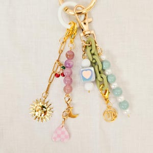 May include: A gold-toned keychain with multiple charms. Charms include a sun, cherries, a moon, a pink heart, a square with a heart, green chain links, and beads in pink, white, and green. The keychain has a white and gold ring.