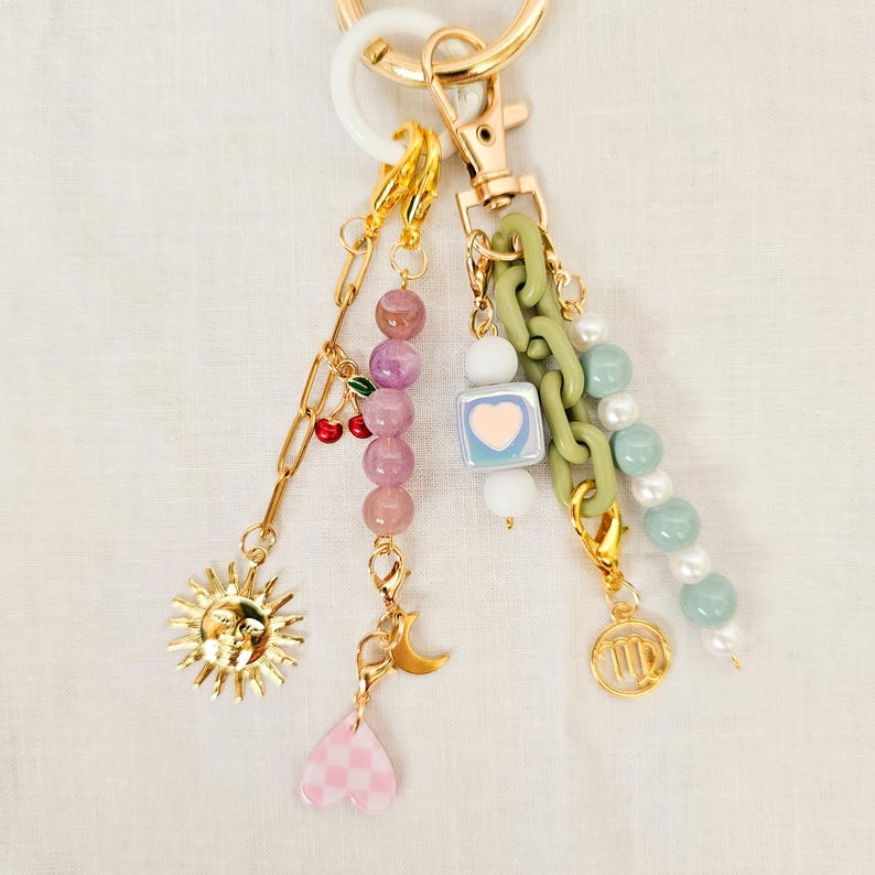 May include: A gold-toned keychain with multiple charms. Charms include a sun, cherries, a moon, a pink heart, a square with a heart, green chain links, and beads in pink, white, and green. The keychain has a white and gold ring.