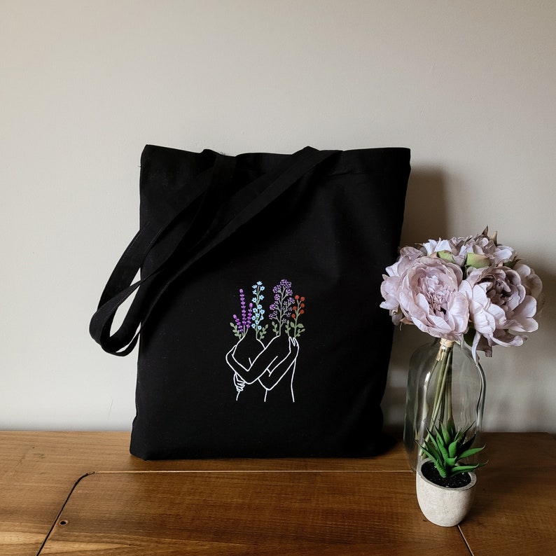 May include: Black tote bag with a white embroidered design of two figures embracing with flowers in their hair. The bag has a long strap and is made of canvas.