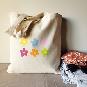 Peut inclure: Un sac fourre-tout en toile beige avec six motifs de fleurs color&eacute;s. Les fleurs sont dans des tons de rose, jaune, bleu et vert.