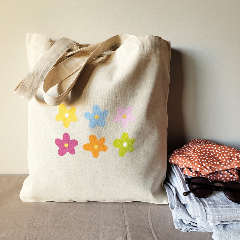 Peut inclure: Un sac fourre-tout en toile beige avec six motifs de fleurs color&eacute;s. Les fleurs sont dans des tons de rose, jaune, bleu et vert.