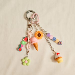 Puede incluir: Un llavero plateado con múltiples dijes. Los dijes incluyen un helado rosa, un girasol, una magdalena, una flor y rodajas de fruta. El llavero también tiene un acento en forma de corazón rosa y una cadena.