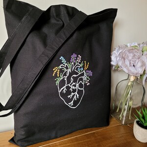 K&ouml;nnte beinhalten: Schwarze Tragetasche mit einem wei&szlig;en gestickten anatomischen Herzen, umgeben von bunten Blumen.