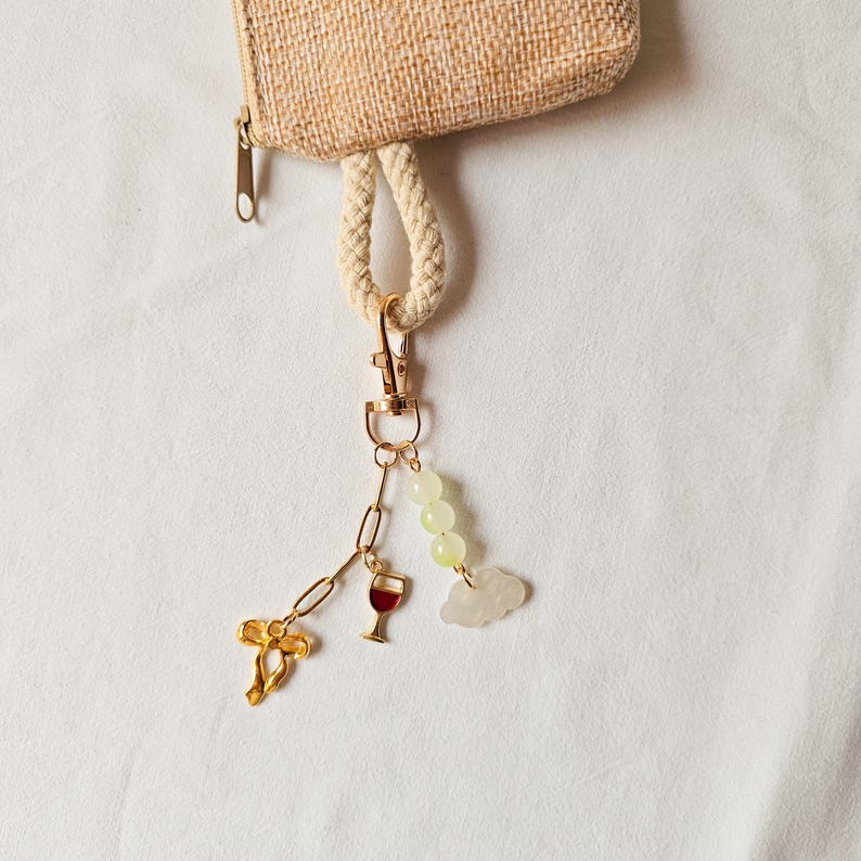 Puede incluir: Un llavero dorado con un dije de copa de vino, un dije de nube y un dije de cuentas verdes. El llavero est&aacute; unido a una peque&ntilde;a bolsa tejida de color beige.