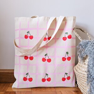 May include: A pink and white canvas tote bag with a checkered pattern and red cherries painted on it. The bag has two handles and is made of natural canvas.