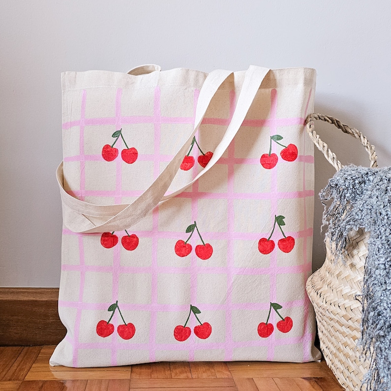 May include: A pink and white canvas tote bag with a checkered pattern and red cherries painted on it. The bag has two handles and is made of natural canvas.
