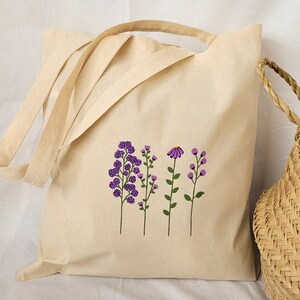 May include: A natural canvas tote bag with a design of purple flowers and green stems. The bag has two long straps and a short handle.