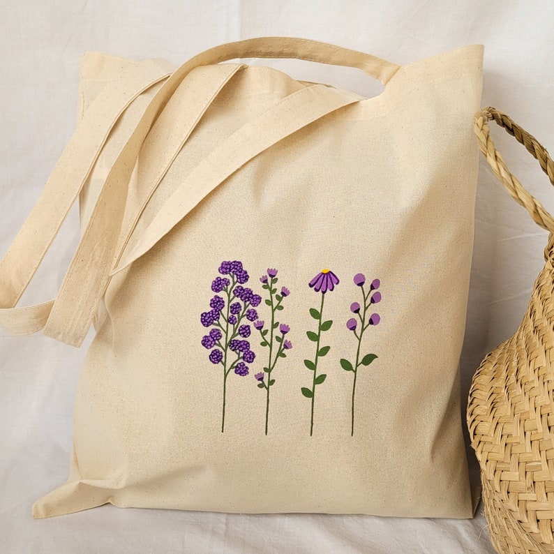 May include: A natural canvas tote bag with a design of purple flowers and green stems. The bag has two long straps and a short handle.