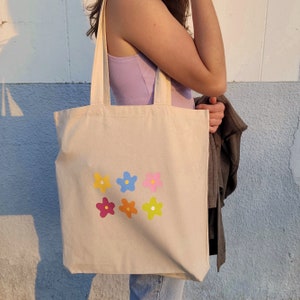 Peut inclure: Un sac fourre-tout en toile beige avec six motifs de fleurs color&eacute;s imprim&eacute;s sur le devant. Les fleurs sont dans des tons de rose, bleu, jaune et orange.