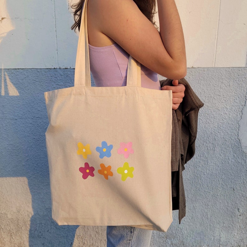 Peut inclure: Un sac fourre-tout en toile beige avec six motifs de fleurs color&eacute;s imprim&eacute;s sur le devant. Les fleurs sont dans des tons de rose, bleu, jaune et orange.