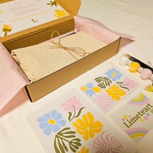 May include: A craft kit in a cardboard box, containing a canvas tote bag, paint, brushes, and floral design templates. The box is lined with pink tissue paper. The kit includes paint in white, yellow, and pink.