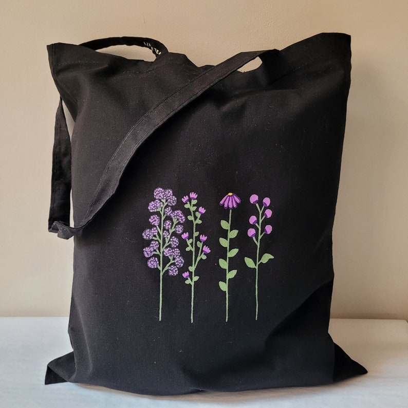 May include: Black tote bag with a floral design embroidered on the front. The flowers are purple and green.