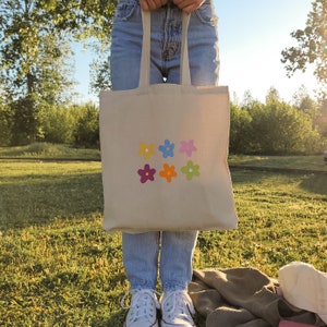 Peut inclure: Un sac fourre-tout beige avec des fleurs brod&eacute;es color&eacute;es. Le sac a deux longues lani&egrave;res et est tenu par une personne.