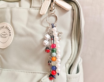 May include: A decorative keychain with a silver ring and lobster claw clasp. The keychain features white dice, red ladybug charms, and a strand of colorful beads, including red, green, orange, and blue. A strand of white pearls is also included.
