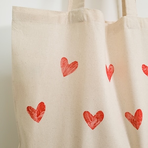 May include: A white canvas tote bag with red heart designs printed on it. The hearts are different sizes and are scattered across the bag.
