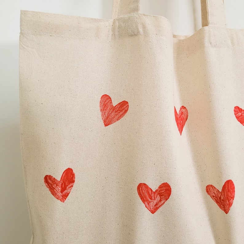 May include: A white canvas tote bag with red heart designs printed on it. The hearts are different sizes and are scattered across the bag.