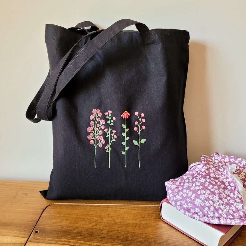 K&ouml;nnte beinhalten: Schwarze Canvas-Tragetasche mit gestickten Blumenmotiven in Rosa und Gr&uuml;n. Die Tasche hat zwei Henkel und ein gro&szlig;es Hauptfach.