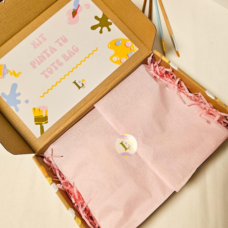May include: An open cardboard box containing a kit for painting a tote bag. The kit includes a card with the text "KIT PINTA TU TOTE BAG" and colorful paint splatters, a pink paper-wrapped item, and pink shredded paper.