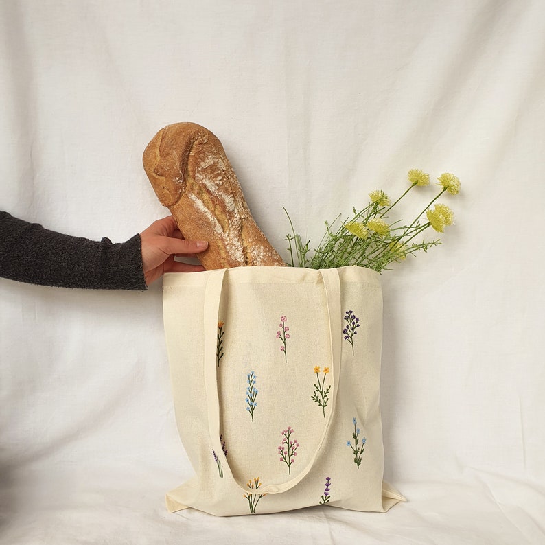 May include: A white canvas tote bag with embroidered flowers in shades of blue, pink, yellow, and purple. A loaf of bread is partially inside the bag.