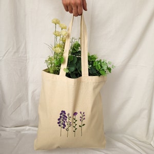 May include: A beige tote bag with a design of purple flowers embroidered on the front. The bag has two straps and is made of canvas.