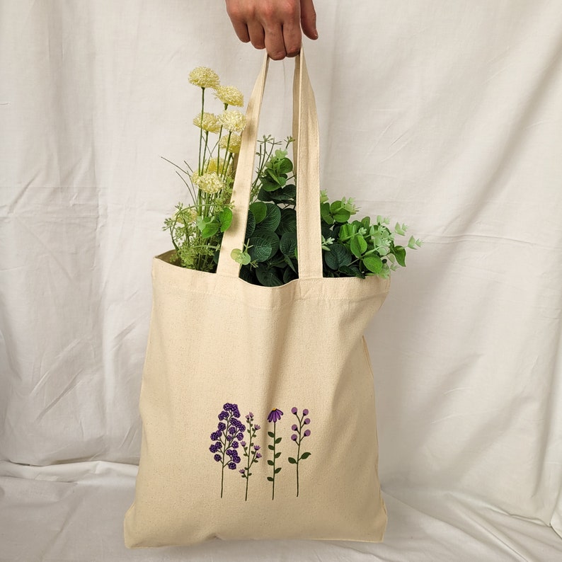 May include: A beige tote bag with a design of purple flowers embroidered on the front. The bag has two straps and is made of canvas.