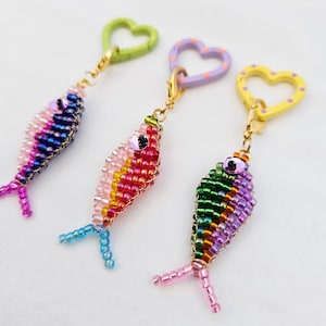 May include: Three beaded fish keychains in shades of pink, blue, green, and purple. Each fish has a different colored heart-shaped clasp. The keychains are made of small, colorful beads and have detailed fins.