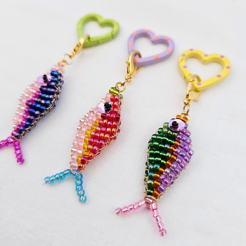 May include: Three beaded fish keychains in shades of pink, blue, green, and purple. Each fish has a different colored heart-shaped clasp. The keychains are made of small, colorful beads and have detailed fins.