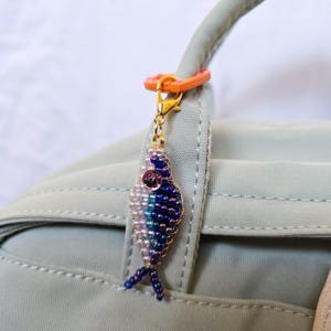 May include: A beaded fish charm in shades of blue, purple, and white, with a gold-colored clasp and orange ring. The charm is attached to a light green bag handle. The fish has a detailed beaded design and a small, pink sequin eye.