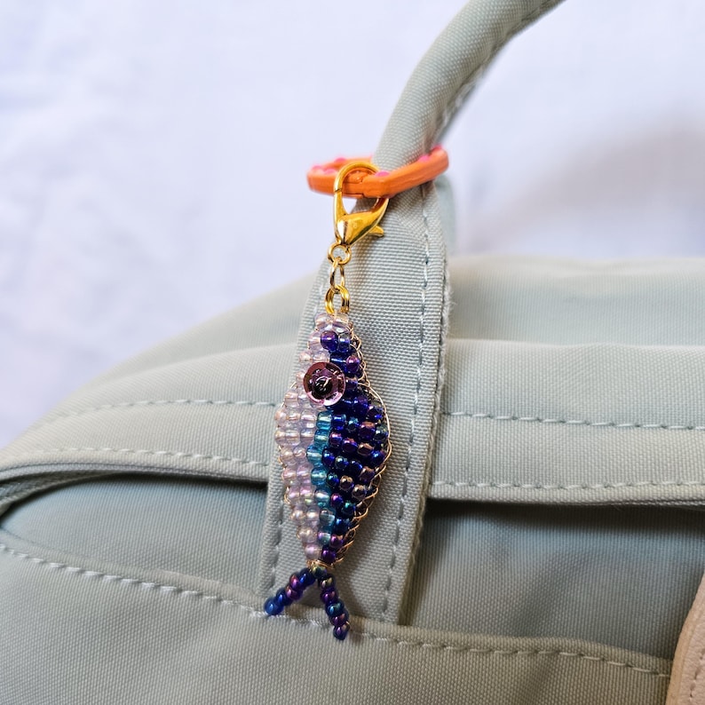 May include: A beaded fish charm in shades of blue, purple, and white, with a gold-colored clasp and orange ring. The charm is attached to a light green bag handle. The fish has a detailed beaded design and a small, pink sequin eye.