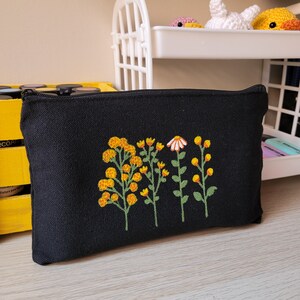 May include: Black canvas zippered pouch with embroidered yellow and white flowers. The flowers are stylized and have a simple design.
