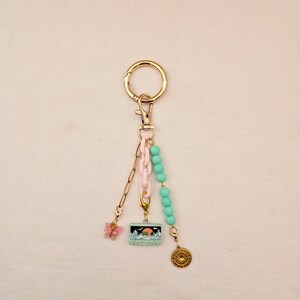 May include: A gold-tone keychain with a lobster clasp, featuring a pink butterfly charm, a small rectangular charm with a mountain scene, and a gold-colored compass charm. The keychain also includes a pink chain and turquoise beads.