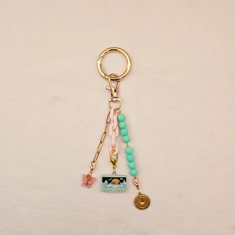 May include: A gold-tone keychain with a lobster clasp, featuring a pink butterfly charm, a small rectangular charm with a mountain scene, and a gold-colored compass charm. The keychain also includes a pink chain and turquoise beads.