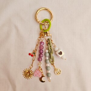 May include: A gold-toned keychain with a green clasp and various charms. Charms include a sun, cherries, a pink heart, a crescent moon, a Gemini symbol, and beaded chains in purple, white, and green. The keychain is designed to hold keys or be used as a decorative accessory.