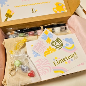 May include: An open cardboard box filled with art supplies. Includes paintbrushes, paint, and decorative items. The box contains a card with the text "Limeteart SHOP" and floral designs. A small bag with a heart charm is visible.